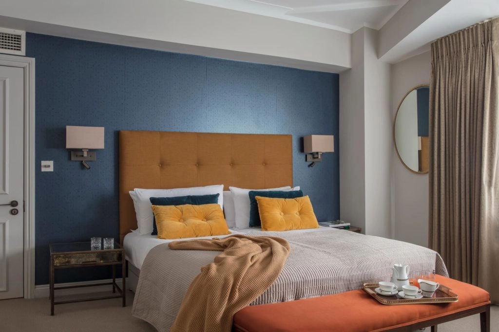 Elegant hotel room with plush bed, blue accent wall, cosy cushions, stylish lighting, and a tea set for a relaxing stay.
