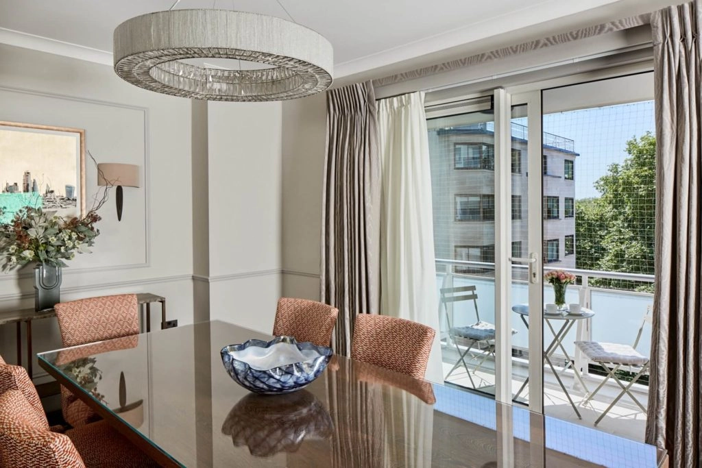 Elegant dining room with a modern chandelier, large table, and balcony access, offering views of lush greenery and stylish patio seating.