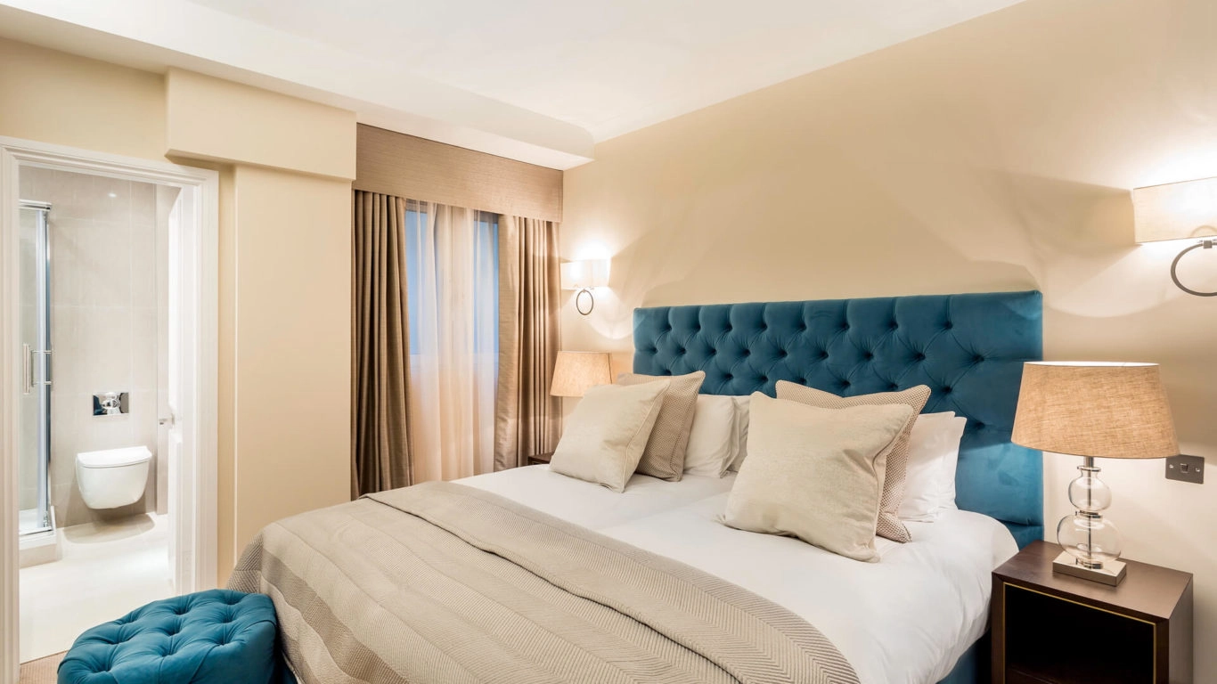 Elegant Arlington House bedroom with plush blue headboard, cosy bedding, warm lighting, and en-suite bathroom.