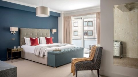 Elegant hotel bedroom with plush bed, vibrant red cushions, chic seating, and large window offering city views at Arlington House Apartments.