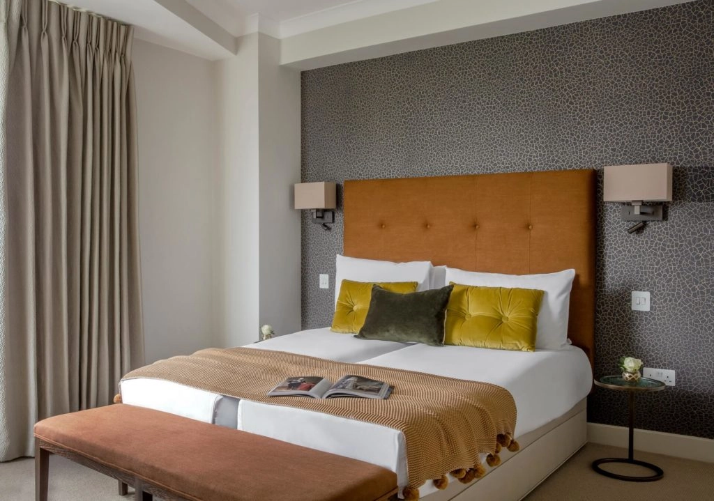 Elegant Arlington House bedroom with plush double bed, textured wallpaper, gold cushions, and modern lighting.