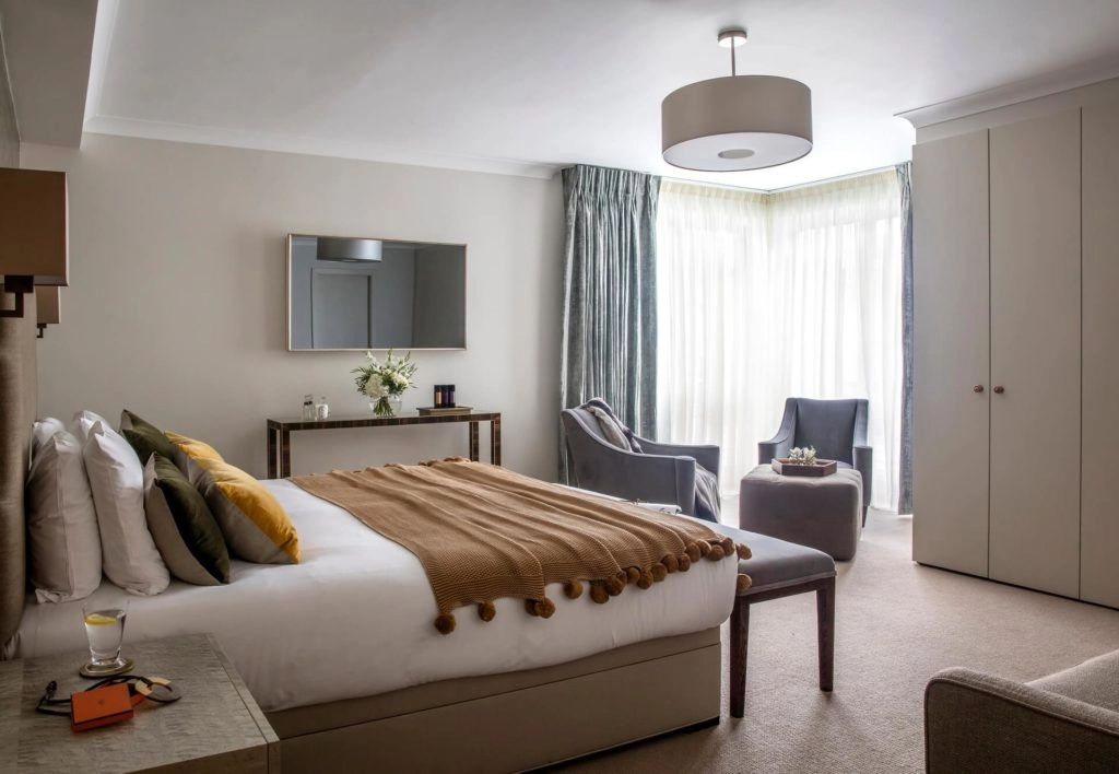 Elegant hotel room with plush bed, cosy seating area, large windows with soft curtains, welcoming atmosphere.