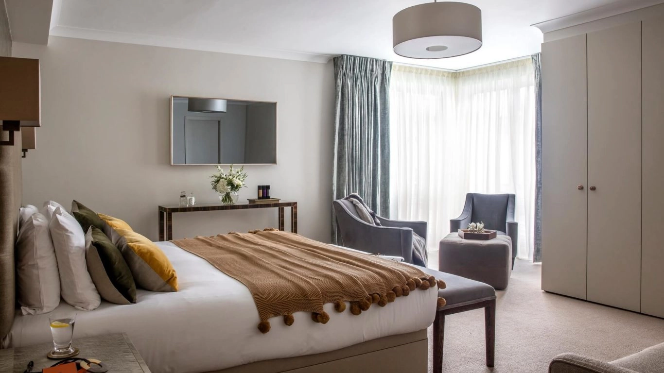 Elegant hotel room with plush bed, cosy seating area, large windows with soft curtains, welcoming atmosphere.