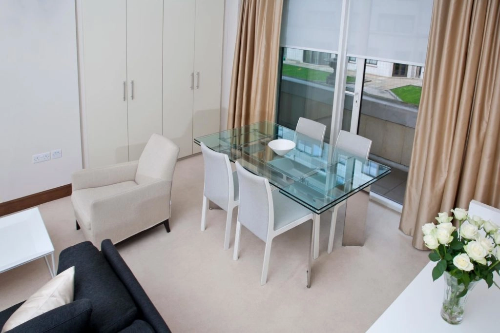 Elegant Dining Area with Roses Elegant dining area with glass table, beige chairs, soft carpeting, and fresh roses, offering comfort and style at Arlington House Apartments.