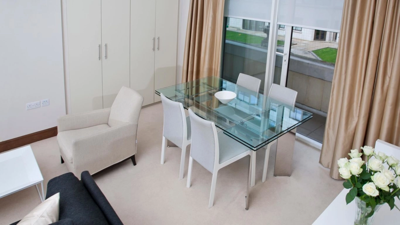 Elegant dining area with glass table, beige chairs, soft carpeting, and fresh roses, offering comfort and style at Arlington House Apartments.